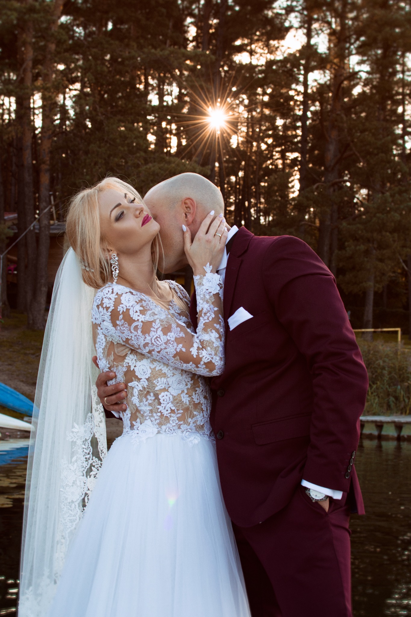 Panna młoda w koronkowej sukni ślubnej i welonie, obejmowana przez pana młodego w bordowym garniturze na tle jeziora i drzew w promieniach zachodzącego słońca.