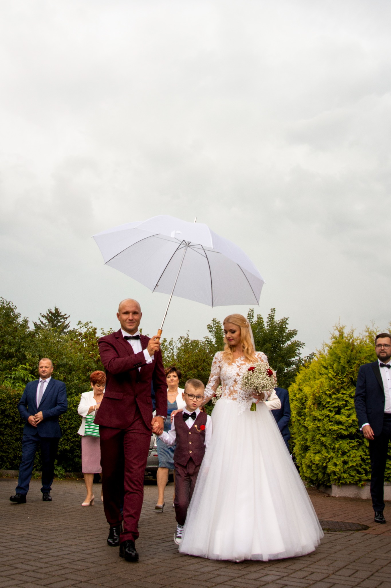 Para młoda w bordowych strojach, on trzyma biały parasol, obok nich idzie chłopiec w garniturze, w tle goście weselni na brukowanej drodze.