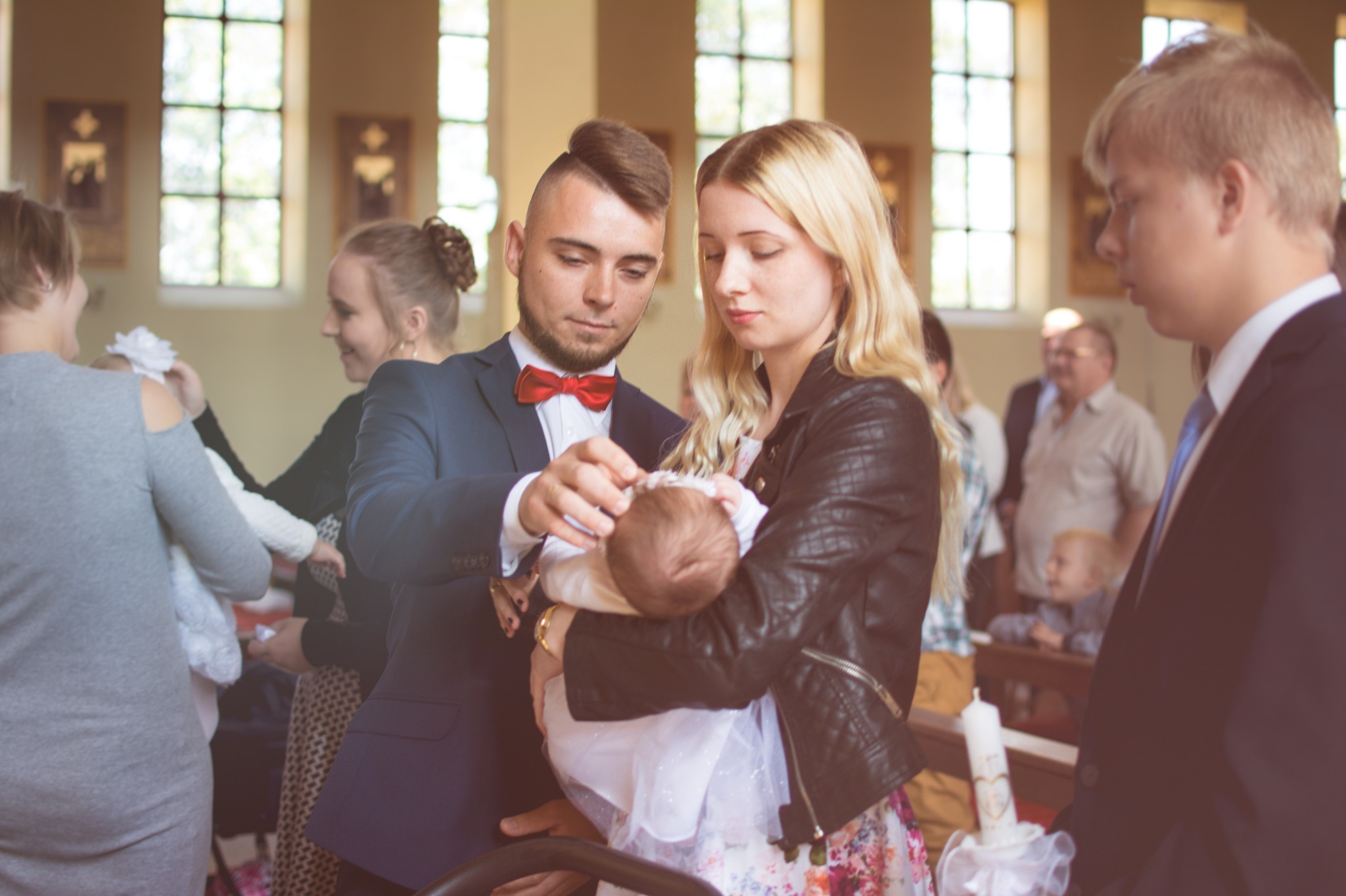 Rodzice w kościele delikatnie poprawiają czapeczkę niemowlęciu ubranemu w białą sukienkę do chrztu, w tle goście i okna