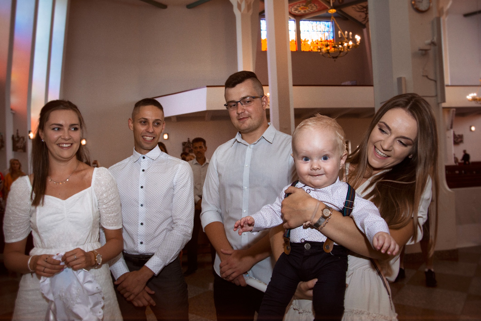 Uśmiechnięta rodzina pozuje w kościele podczas uroczystości, kobieta trzyma na rękach roześmianego chłopca w eleganckich spodniach na szelkach i koszuli.