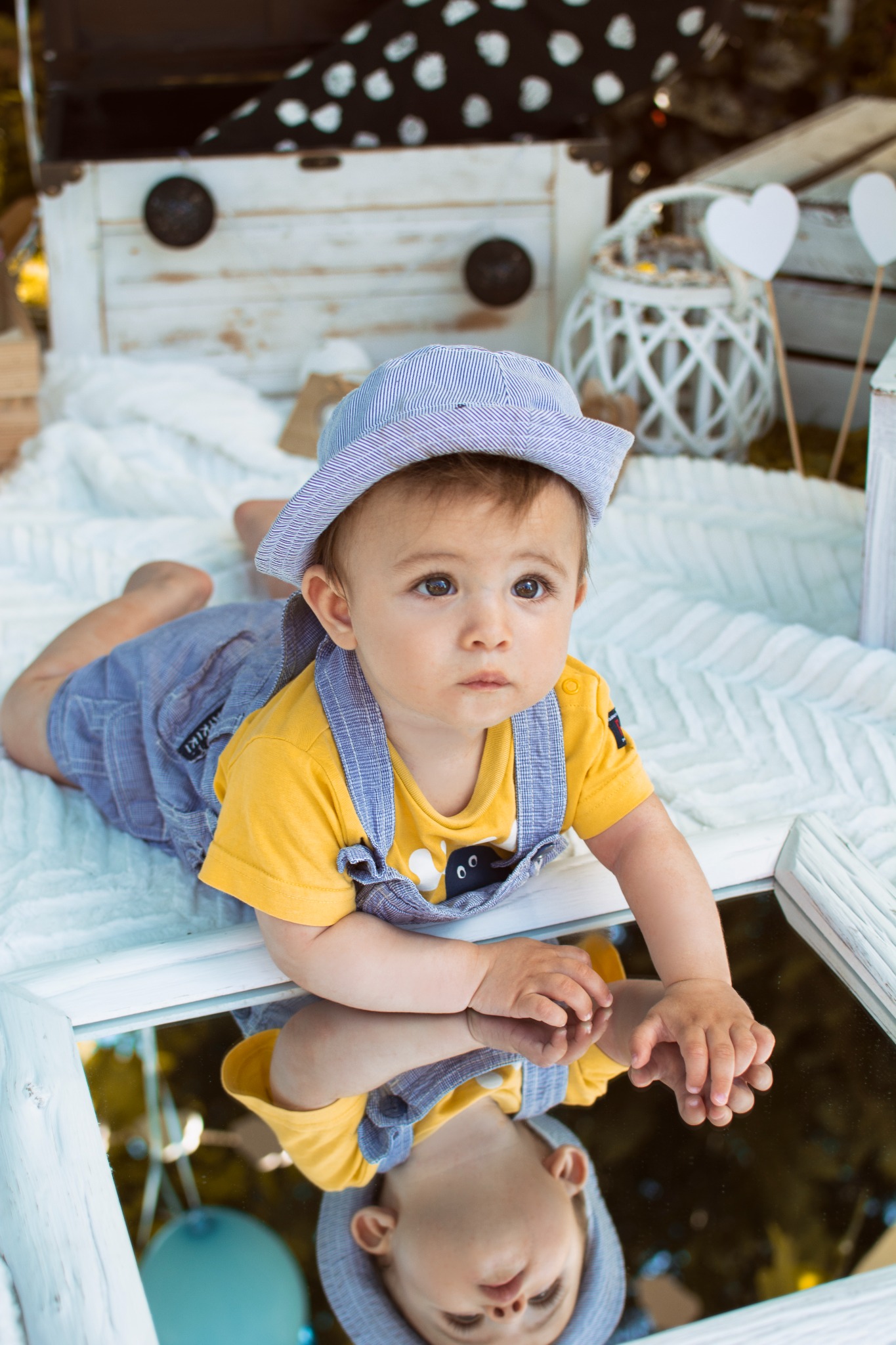 Uroczy niemowlak w paski kapeluszu i żółtej koszulce leży na brzuchu, opierając dłonie na lustrzanym blacie, z odbiciem w lustrze, w tle białe skrzynie i dekoracje.