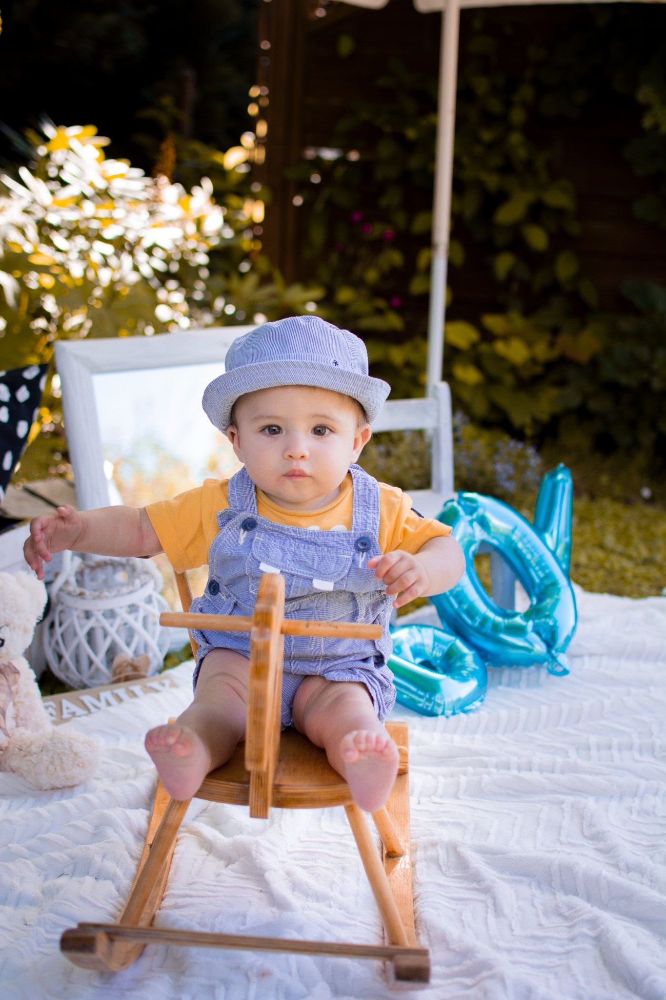 Uroczy niemowlak w niebieskim kapeluszu i ogrodniczkach siedzi na drewnianym koniku na biegunach podczas sesji zdjęciowej na białym kocu z dekoracjami, w tle zieleń i biała rama.
