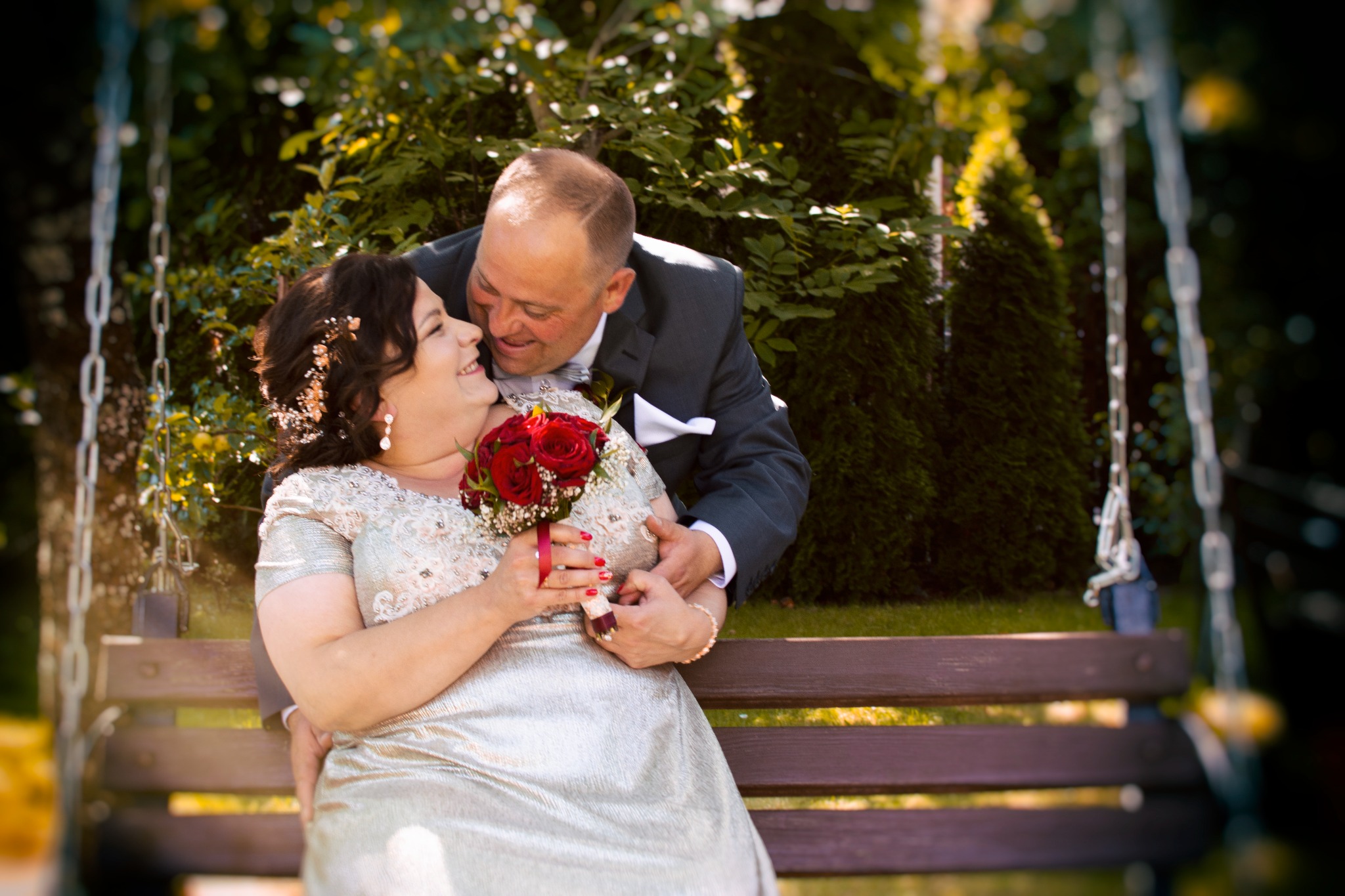 Uśmiechnięta para młoda siedząca na huśtawce ogrodowej, panna młoda trzyma bukiet czerwonych róż, a pan młody obejmuje ją czule.