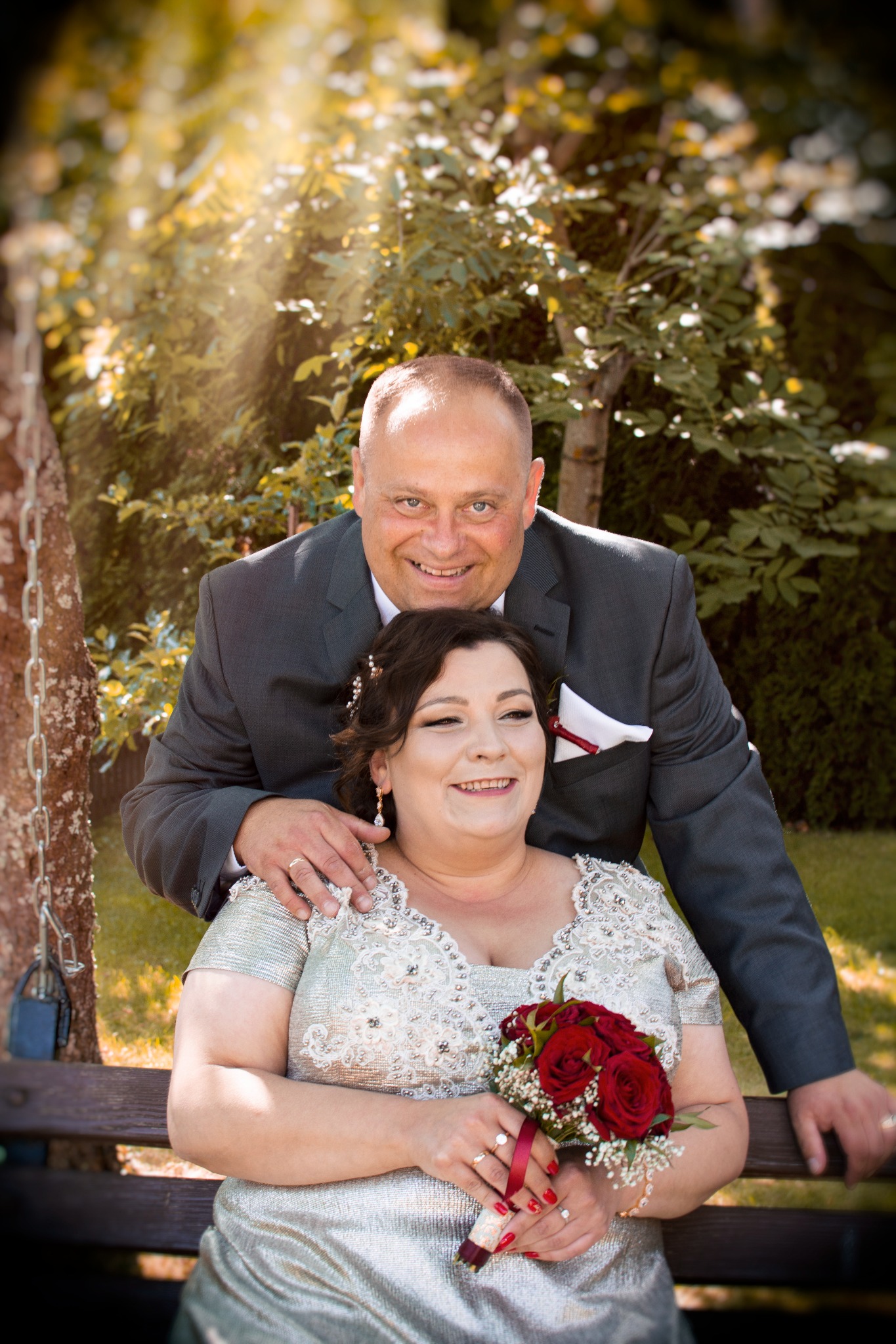 Uśmiechnięta para siedząca na drewnianej huśtawce w ogrodzie: kobieta w srebrnej sukni z bukietem czerwonych róż, mężczyzna w szarym garniturze z butonierką.