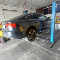 Ciemny samochód sedan na podnośniku w warsztacie, widok z tyłu, z widocznymi złotymi felgami i elementami oświetlenia warsztatowego na suficie.