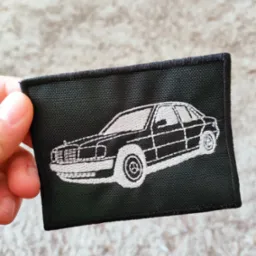 Czarna naszywka z wyhaftowanym białym konturem samochodu Mercedes W124 trzymana w dłoni na tle puszystego dywanu.