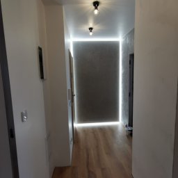 Smart Interiors - Korytarz z drewnianą podłogą, szarą ścianą z efektem betonu, oświetleniem LED wzdłuż krawędzi ściany i dwoma czarnymi lampami sufitowymi.