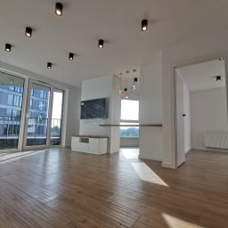 Smart Interiors - Jasne, przestronne wnętrze mieszkania z drewnopodobną podłogą, białymi ścianami, czarnymi reflektorami sufitowymi i dużymi oknami balkonowymi. Telewizor zabezpieczony folią na ścianie, biała szafka...