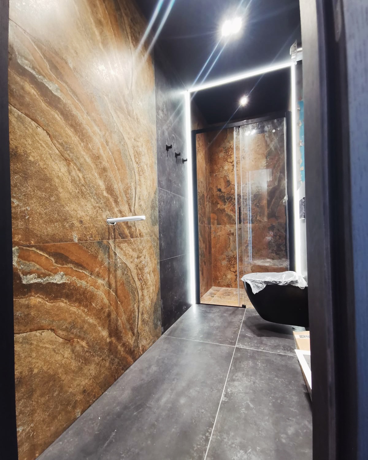 Nowoczesna łazienka z brązowymi płytkami imitującymi kamień, czarną armaturą i podświetleniem LED wokół kabiny prysznicowej. Wanna wolnostojąca zabezpieczona folią ochronną.