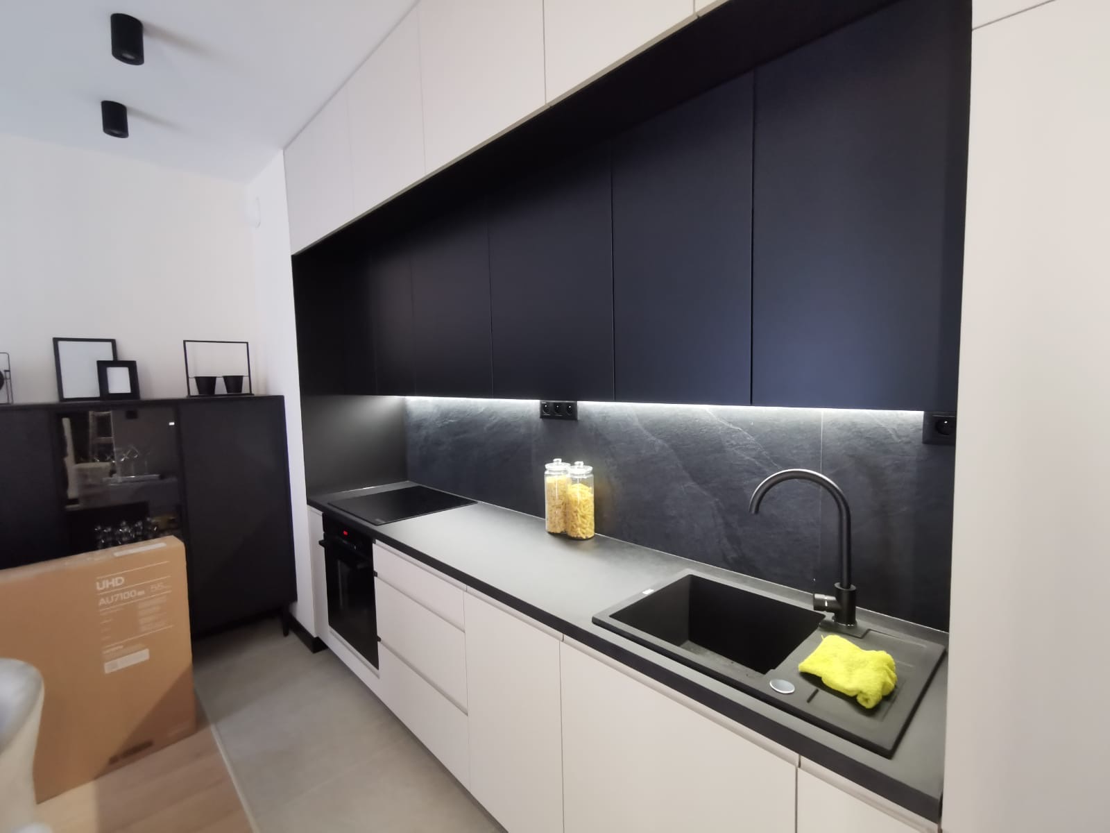 Nowoczesna kuchnia z białymi szafkami dolnymi, czarnymi szafkami górnymi i blatem, czarnym zlewozmywakiem i baterią, podświetleniem LED pod szafkami, oraz dekoracyjnym panelem ściennym imitującym...