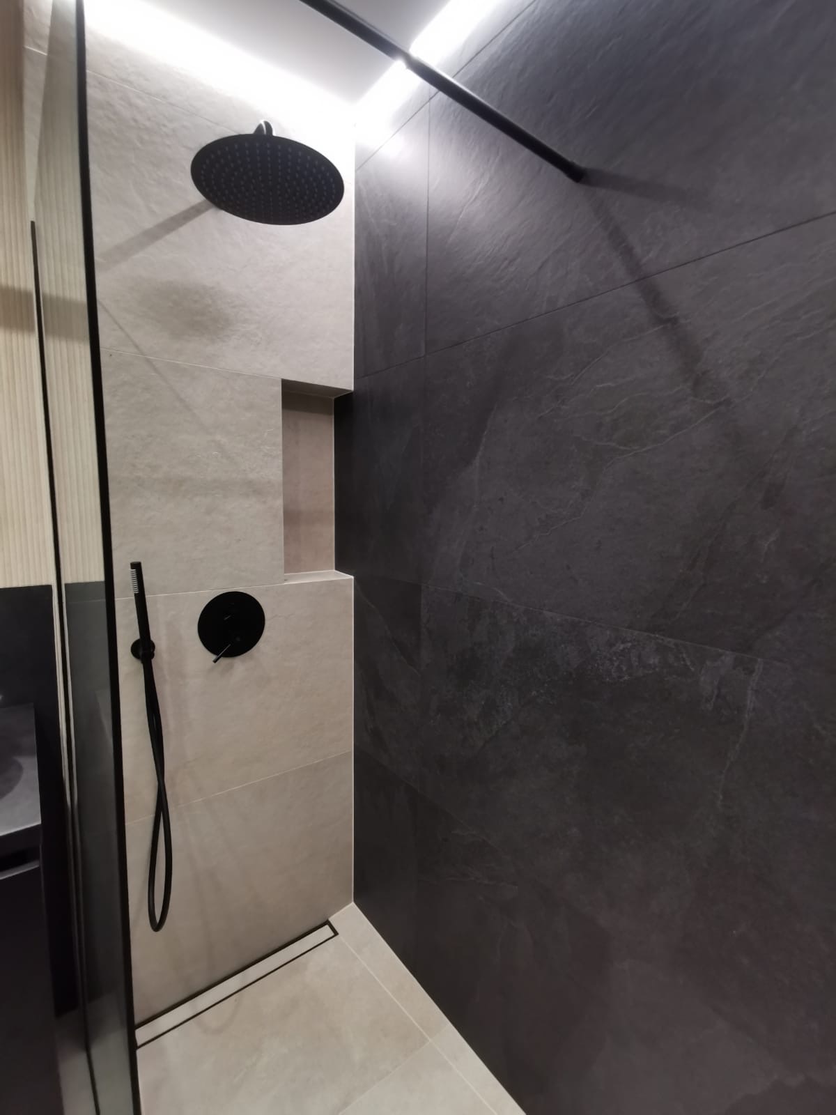 Nowoczesna kabina prysznicowa walk-in z czarną armaturą, jasnymi płytkami i ciemnoszarą ścianą z efektem kamienia.