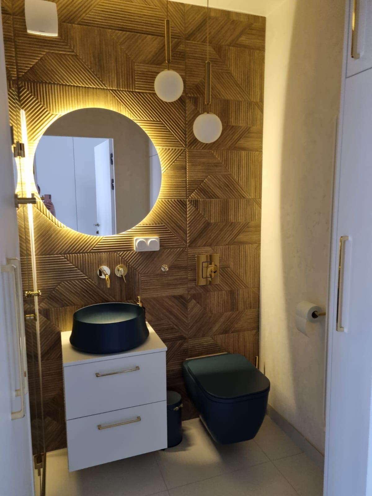 Nowoczesna łazienka z drewnianą ścianą w geometryczne wzory, okrągłym lustrem z podświetleniem LED, ciemnoniebieską umywalką nablatową i toaletą w tym samym kolorze, białą szafką pod umywalką...