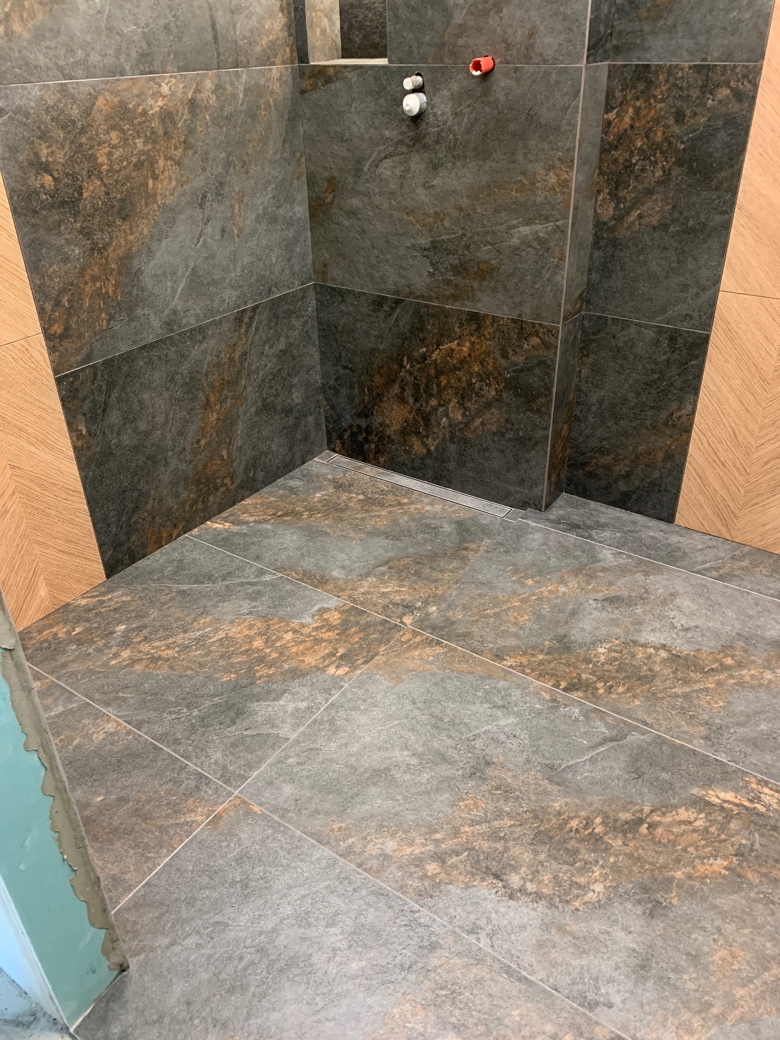 Ukończona kabina prysznicowa z szarymi płytkami imitującymi kamień, z widocznym odpływem liniowym i fragmentem drewnopodobnej okładziny na ścianie.