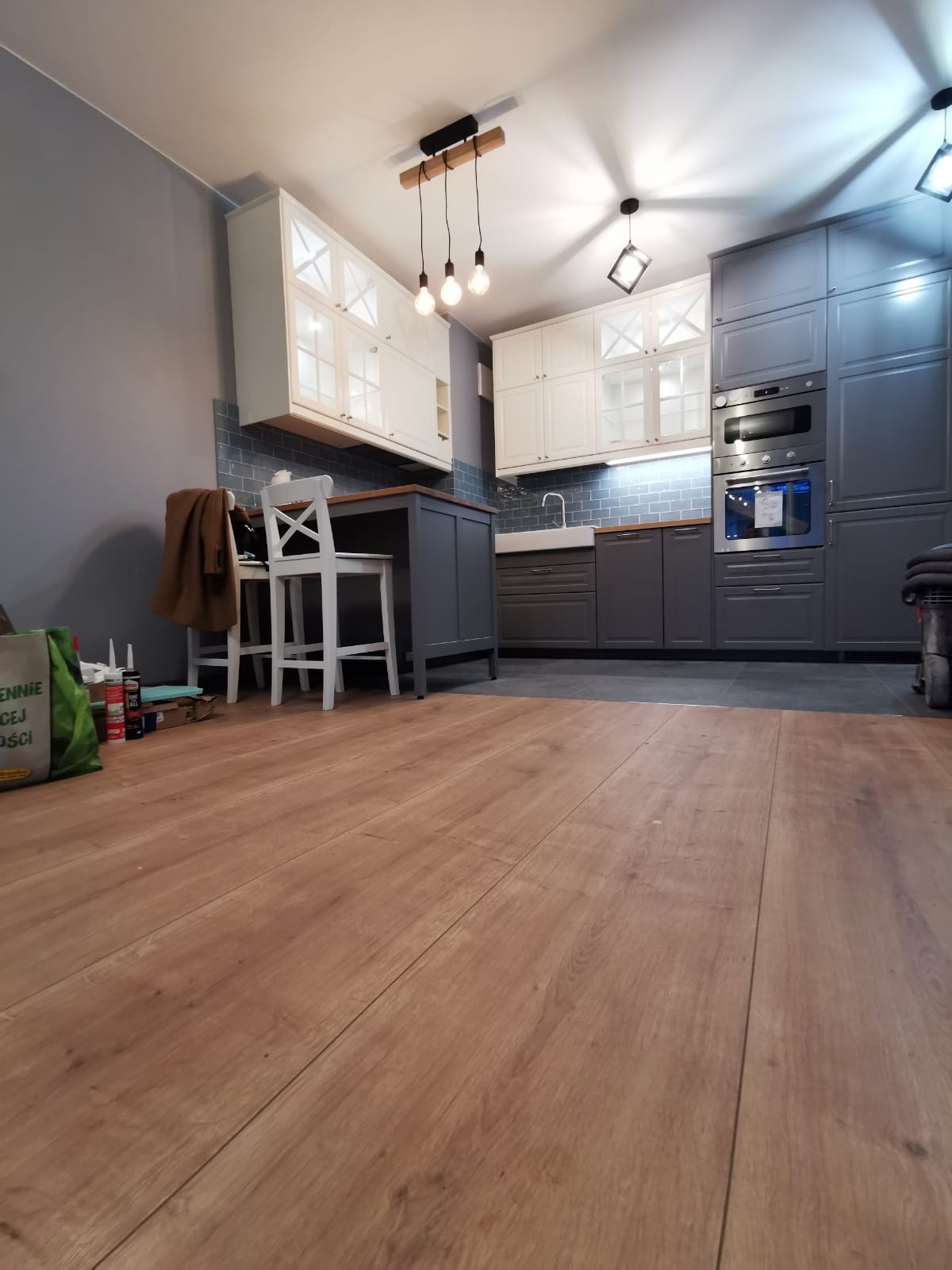 Nowoczesna kuchnia w odcieniach szarości i bieli z drewnianym blatem, widok z poziomu podłogi na panele, oświetlenie punktowe i wiszące żarówki.