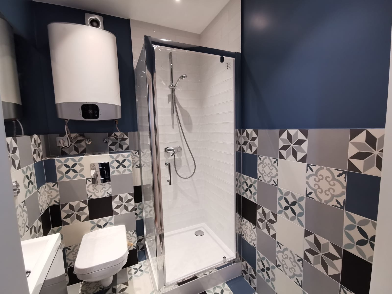 Nowoczesna łazienka z kabiną prysznicową, wiszącą toaletą i wzorzystymi płytkami w odcieniach szarości i błękitu, z widocznym bojlerem elektrycznym na ścianie.