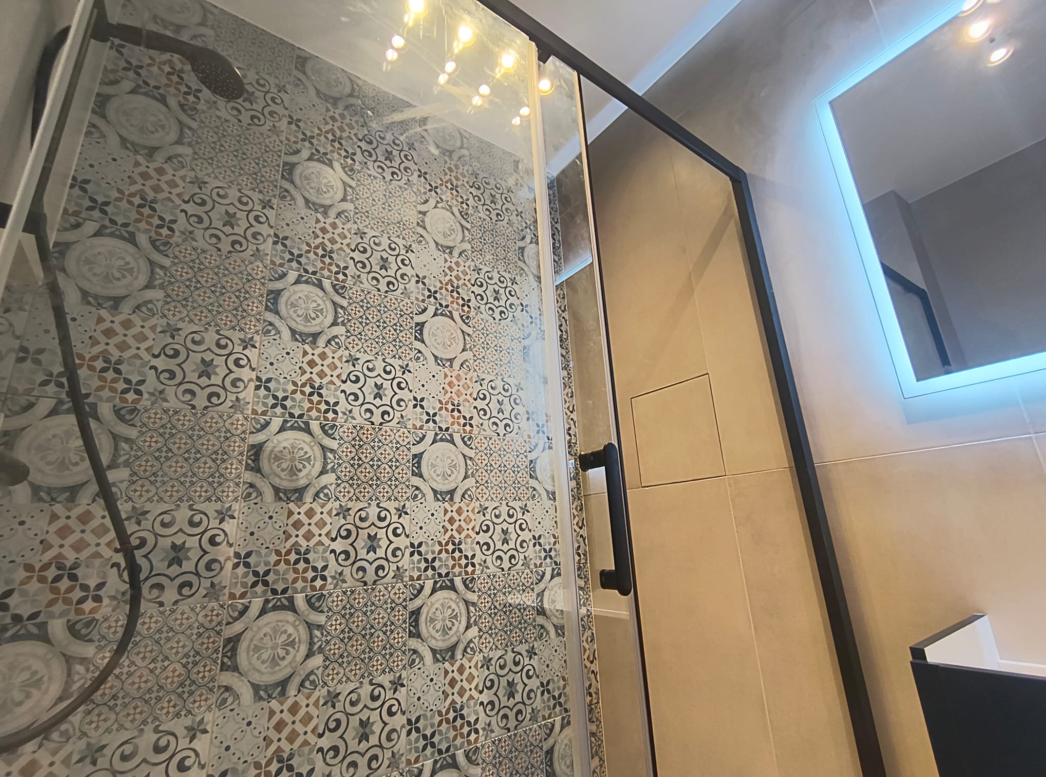 Nowoczesna kabina prysznicowa z czarnymi profilami i drzwiami, ściana wyłożona dekoracyjnymi płytkami patchworkowymi, lustro z podświetleniem LED.