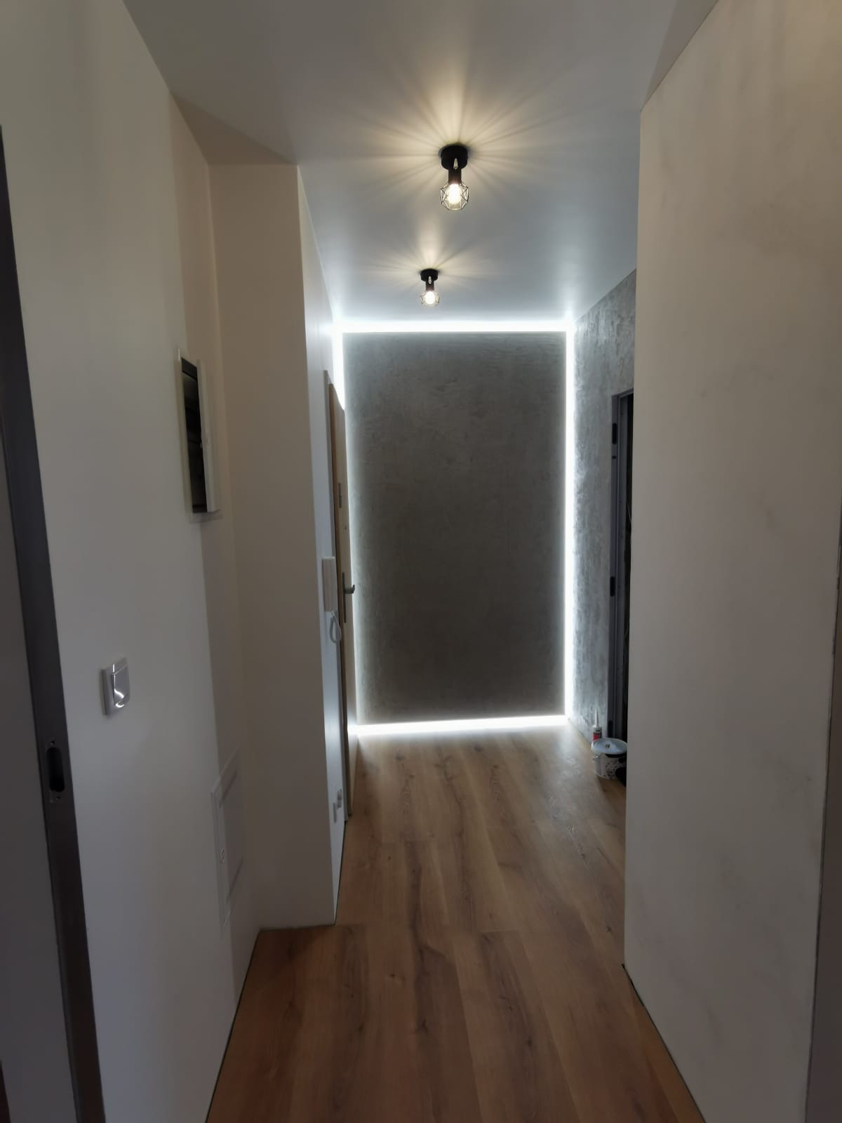 Korytarz z drewnianą podłogą, szarą ścianą z efektem betonu, oświetleniem LED wzdłuż krawędzi ściany i dwoma czarnymi lampami sufitowymi.