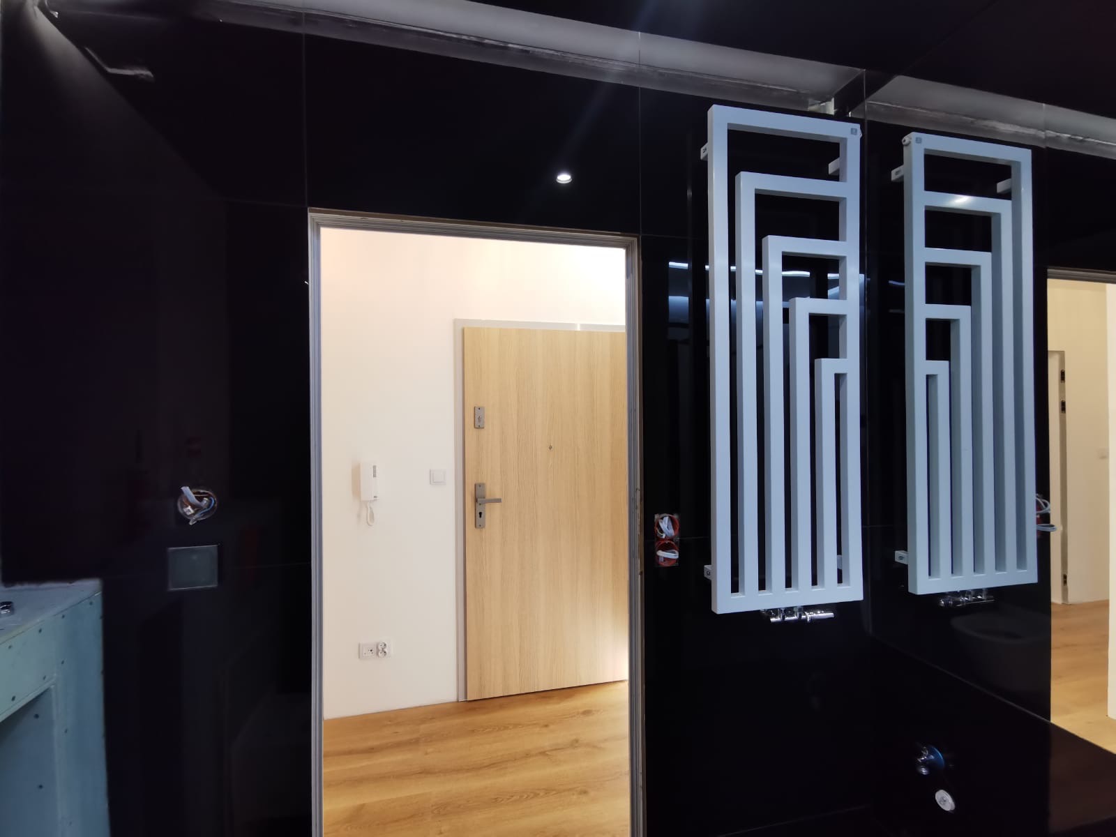 Czarne, błyszczące ściany łazienki z dekoracyjnymi, białymi grzejnikami drabinkowymi, widok na przedpokój z drewnianymi drzwiami wejściowymi.