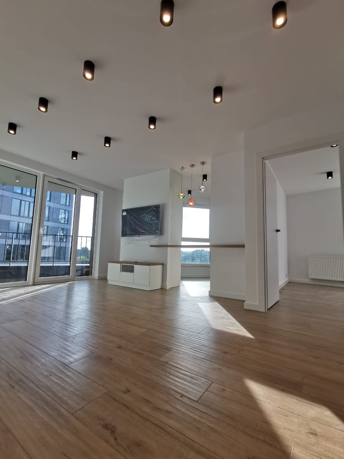 Jasne, przestronne wnętrze mieszkania z drewnopodobną podłogą, białymi ścianami, czarnymi reflektorami sufitowymi i dużymi oknami balkonowymi. Telewizor zabezpieczony folią na ścianie, biała szafka...