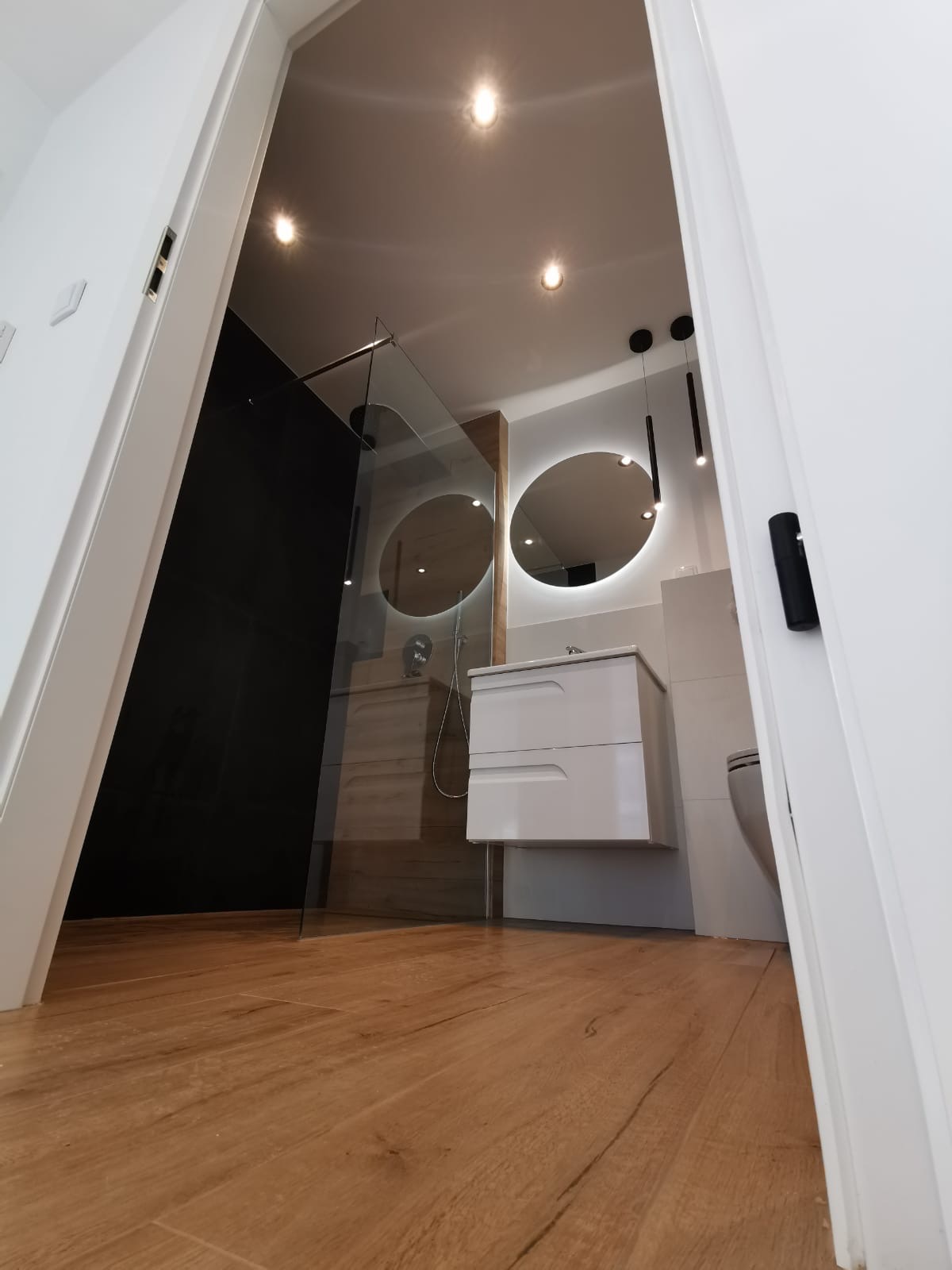 Nowoczesna łazienka z drewnopodobną podłogą, kabiną prysznicową typu walk-in z czarną ścianą, okrągłymi lustrami z podświetleniem LED i białą szafką z umywalką, widziana z perspektywy otwartych drzwi.