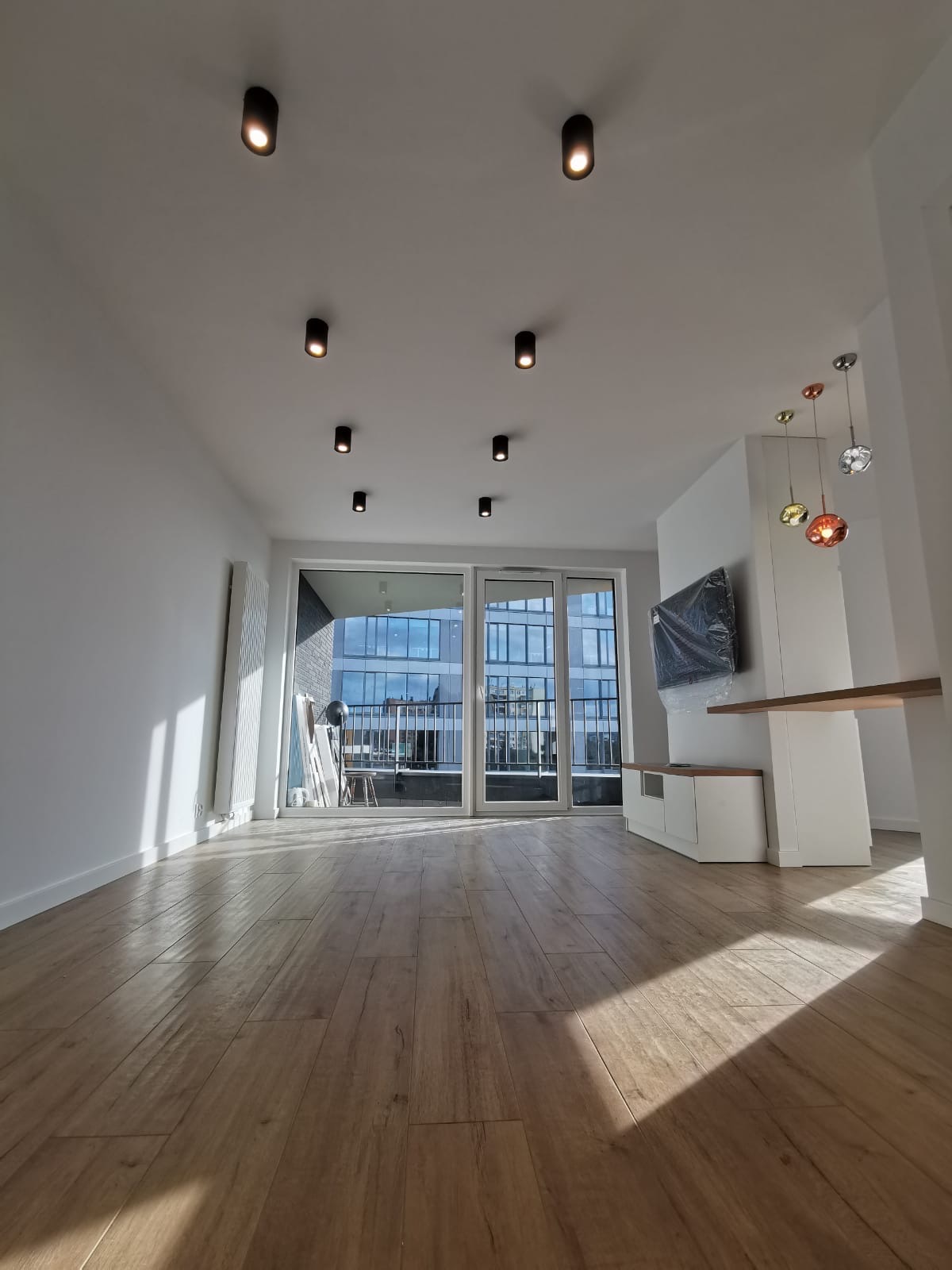 Jasne, przestronne wnętrze po remoncie z drewnopodobną podłogą, białymi ścianami, dużymi oknami balkonowymi i nowoczesnym oświetleniem punktowym na suficie. Telewizor zabezpieczony folią. Widok...