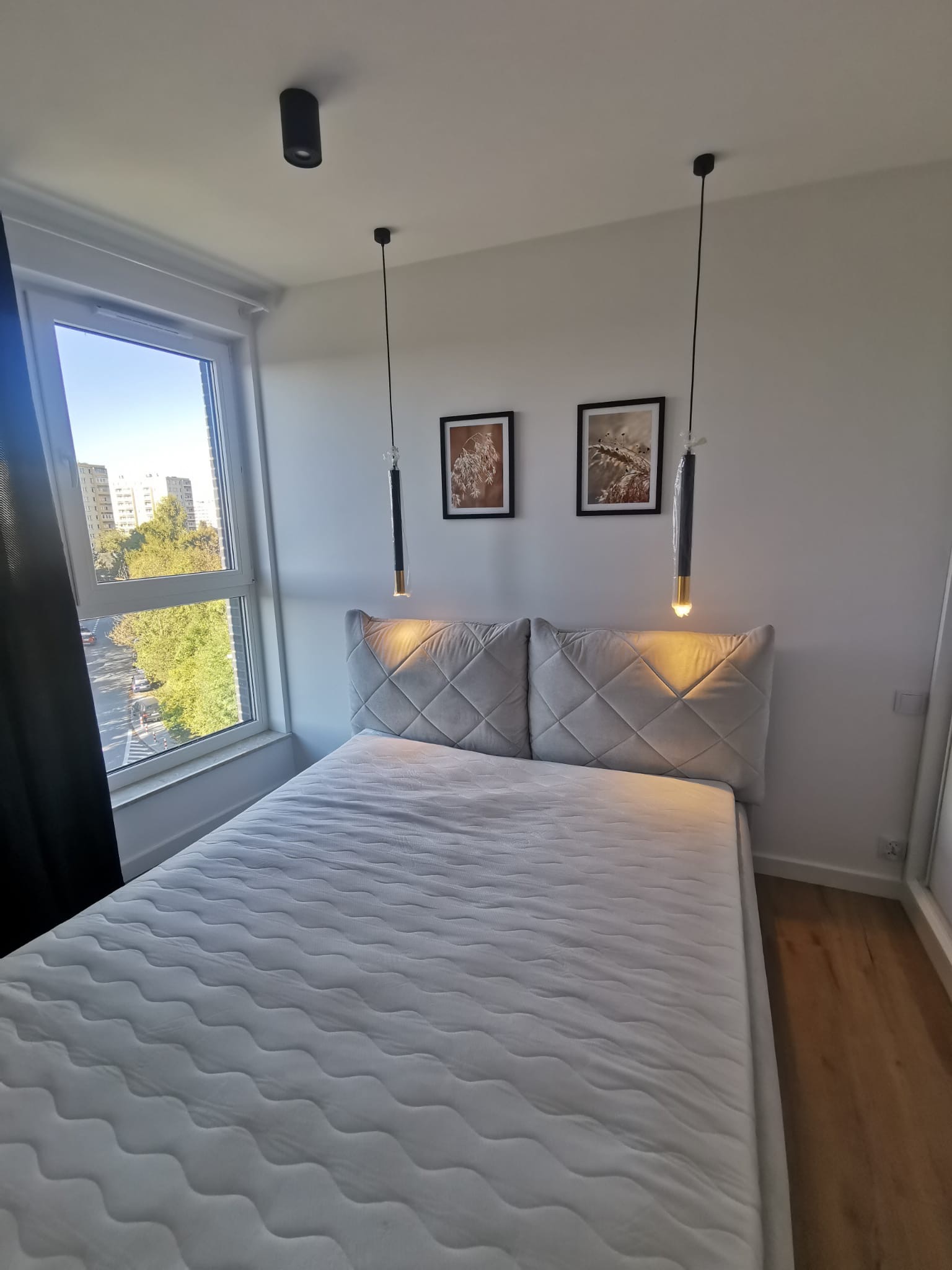 Jasne wnętrze sypialni z łóżkiem z szarym zagłówkiem w romby, wiszącymi lampami z elementami złota i widokiem na miasto z okna.