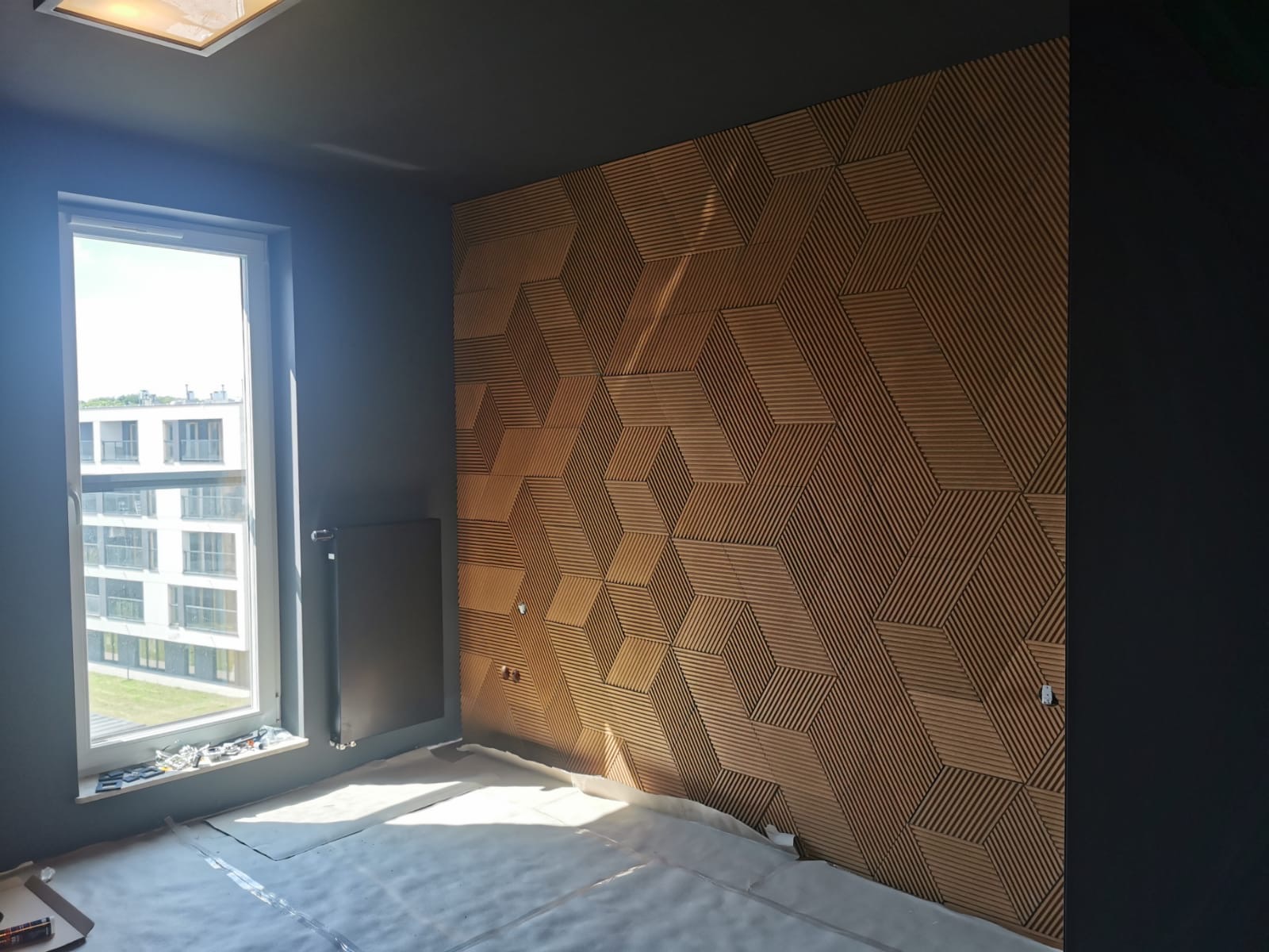 Montaż dekoracyjnej ściany z drewnianych paneli 3D w geometryczne wzory, widok na okno w nowym mieszkaniu w Krakowie, podłoga zabezpieczona folią ochronną.