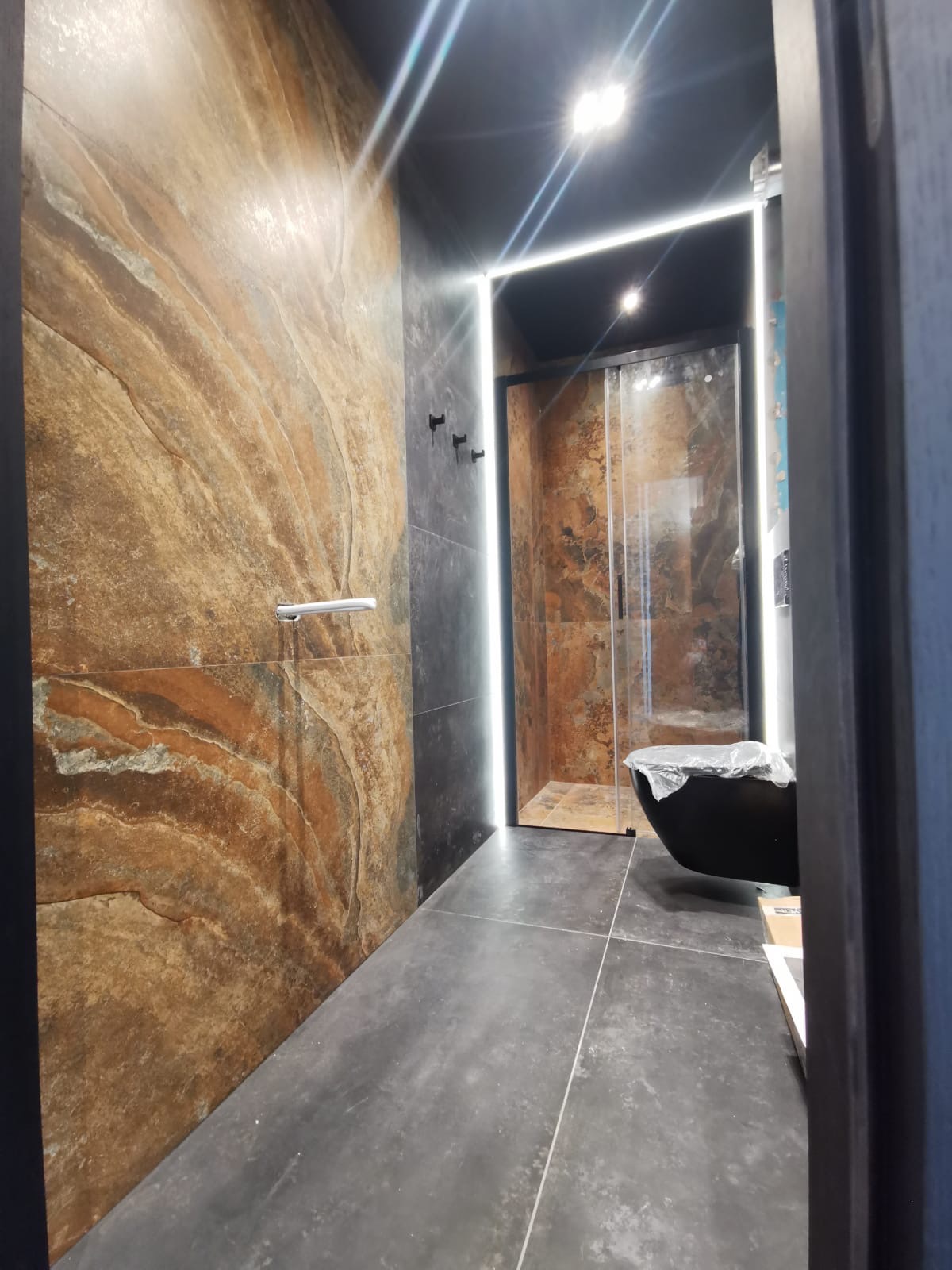 Nowoczesna łazienka z płytkami imitującymi kamień w odcieniach brązu i szarości, podświetlana listwa LED, czarna armatura i kabina prysznicowa. Wanna wolnostojąca przykryta folią ochronną.