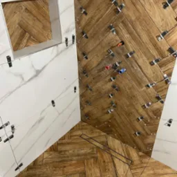 Układanie płytek imitujących drewno w jodełkę na podłodze i marmurowych płytek na ścianie w łazience, z widocznymi krzyżykami dystansowymi i odpływem liniowym.