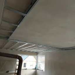 Metalowa konstrukcja sufitu podwieszanego z zamontowaną płytą, widok z dołu, w tle okno z widokiem na dom i drzewa.