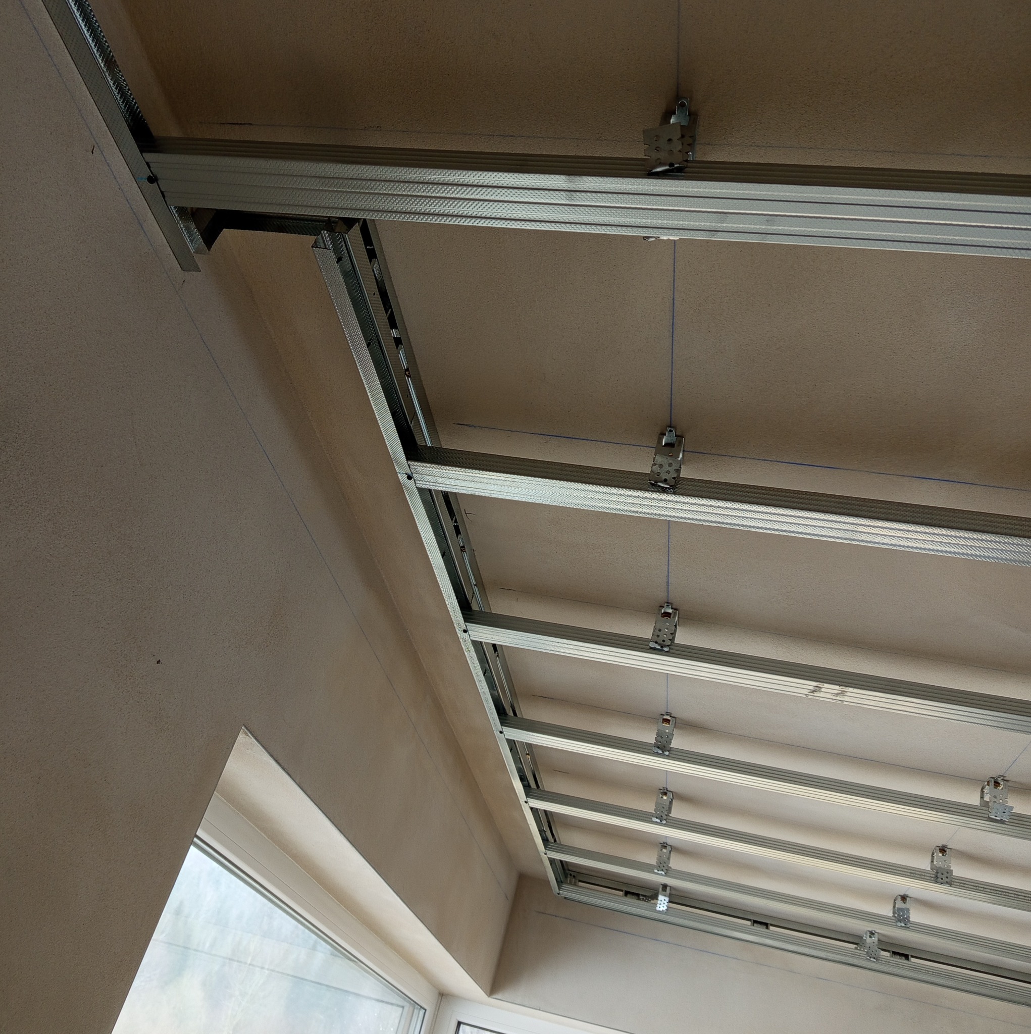 Metalowa konstrukcja sufitu podwieszanego z widocznymi profilami CD i UD, zawiesiami oraz sznurkami poziomującymi, na tle surowego stropu i ściany w trakcie remontu, ujęcie z dołu.