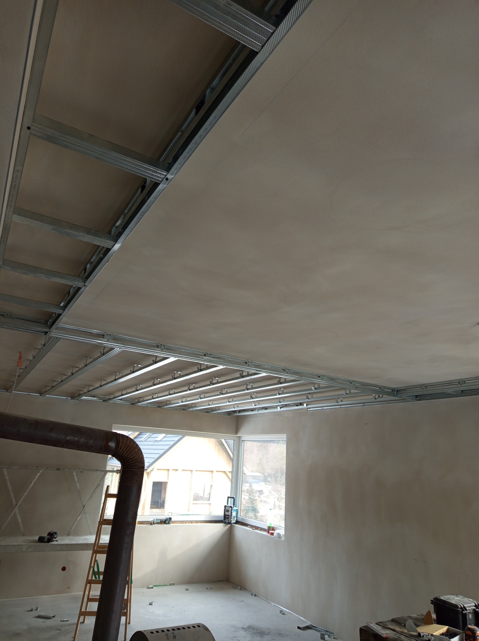 Metalowa konstrukcja sufitu podwieszanego z zamontowaną płytą, widok z dołu, w tle okno z widokiem na dom i drzewa.