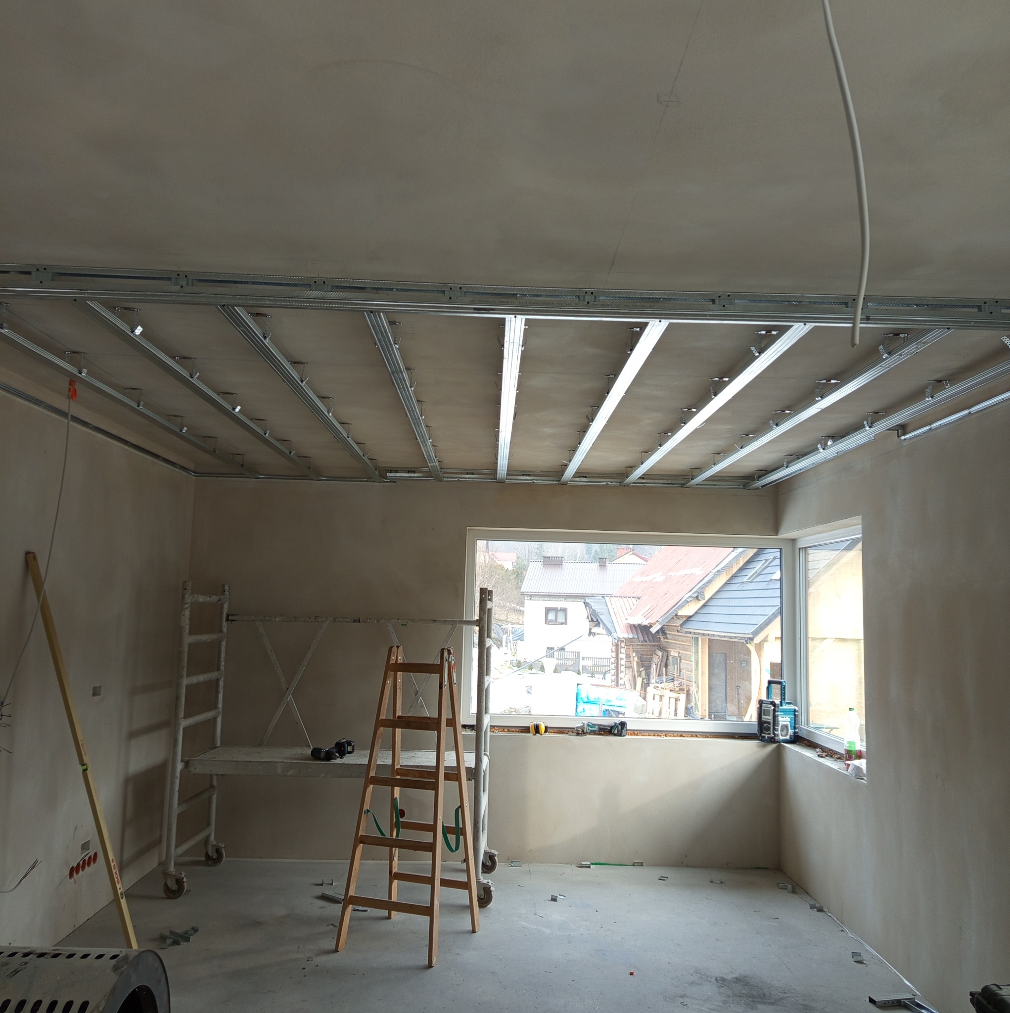 Wnętrze w trakcie prac wykończeniowych: widoczna konstrukcja metalowa podwieszanego sufitu, ściany po wstępnym wykończeniu, drabina i rusztowanie w pomieszczeniu z oknem.