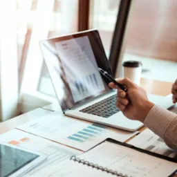 Analiza raportu na laptopie z użyciem długopisu, dokumenty i tablet na biurku, kubek z kawą w tle.