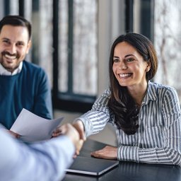 CENTRUM KSIĘGOWO-EKONOMICZNE LUB Sp. z o.o. - Uśmiechnięta kobieta w koszuli w paski wita się z klientem, w tle mężczyzna z dokumentami, spotkanie biznesowe w jasnym biurze.