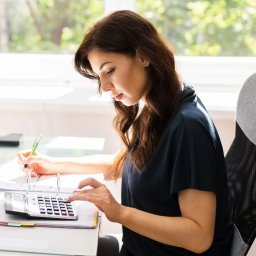 CENTRUM KSIĘGOWO-EKONOMICZNE LUB Sp. z o.o. - Kobieta przy biurku, wpisuje dane do kalkulatora i notuje długopisem na papierach w segregatorze, jasne światło z okna w tle.