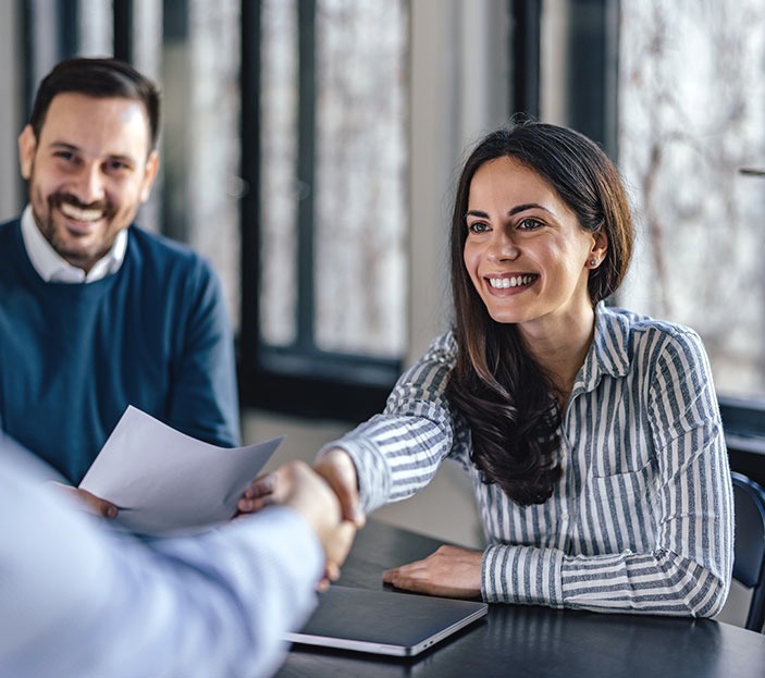 Uśmiechnięta kobieta w koszuli w paski wita się z klientem, w tle mężczyzna z dokumentami, spotkanie biznesowe w jasnym biurze.