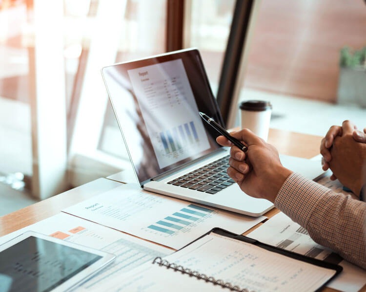 Analiza raportu na laptopie z wykresem słupkowym, dokumenty, notatnik i kawa na biurku w jasnym biurze.