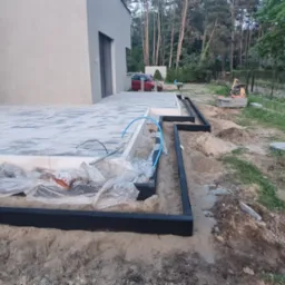 Przygotowanie podłoża pod taras z kostki brukowej: widoczne obrzeża, piasek, folia ochronna, oraz częściowo ułożona kostka na betonowej płycie, w tle dom z szarą elewacją i drzewa.