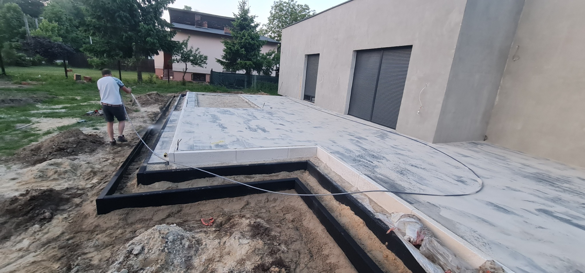 Pracownik spryskuje wodą świeżo ułożoną kostkę brukową na tarasie nowoczesnego domu, widoczne czarne obrzeża i przygotowane podłoże z piasku.