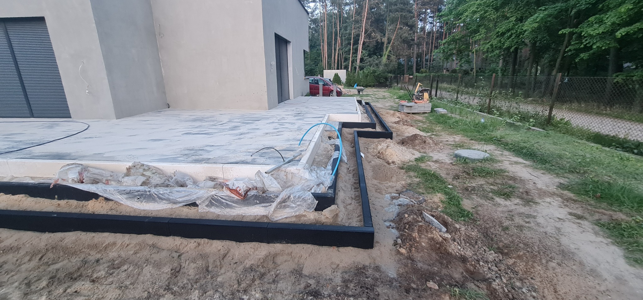 Przygotowanie podłoża pod taras z kostki brukowej: widoczne obrzeża, piasek, folia ochronna, oraz częściowo ułożona kostka na betonowej płycie, w tle dom z szarą elewacją i drzewa.