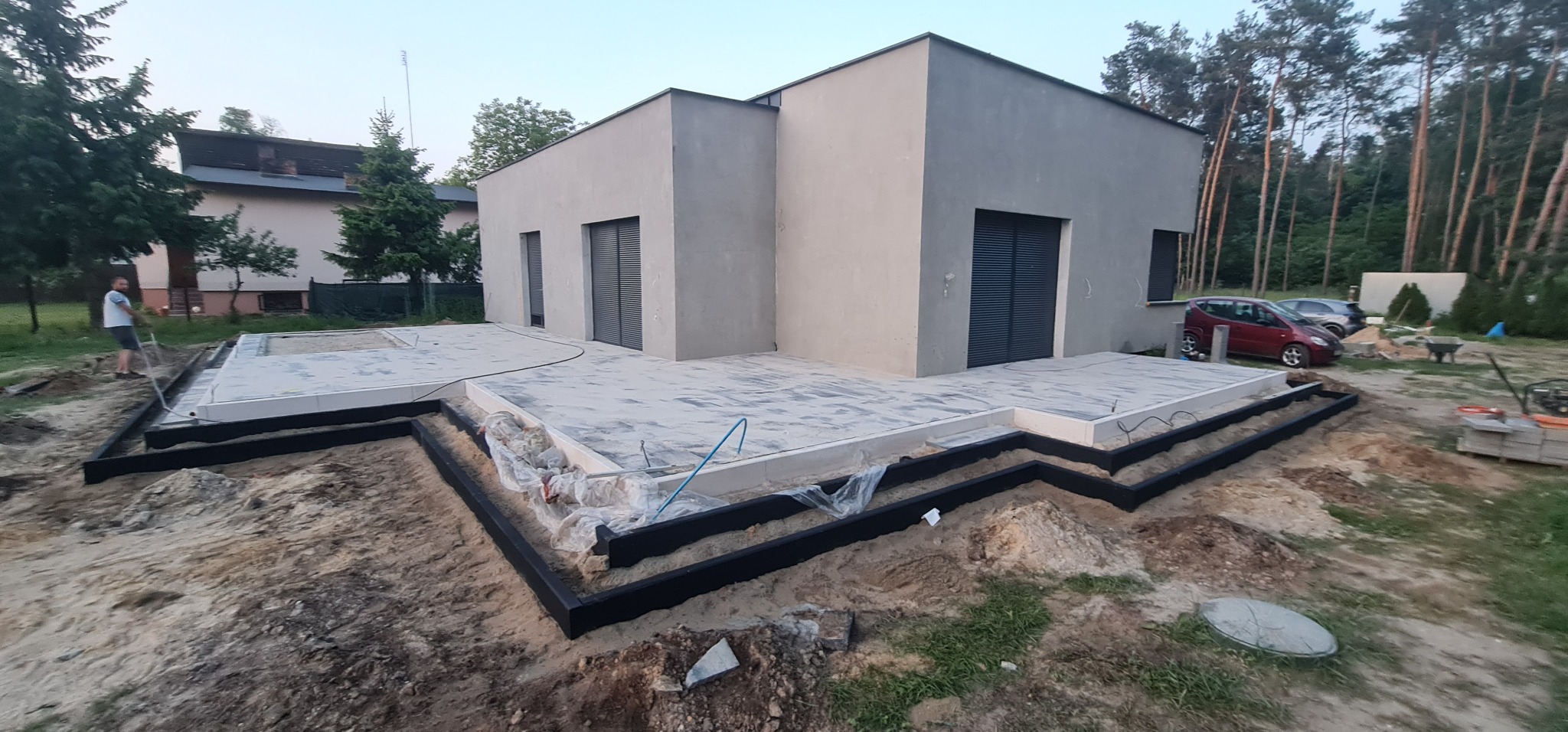 Przygotowanie podłoża pod taras z kostki brukowej: widoczne czarne obrzeża i częściowo ułożone płyty tarasowe na tle nowoczesnego domu z szarymi elewacjami i zielenią drzew