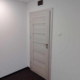 Andrzej Szelag - Wnętrze biura z jasnymi ścianami, ciemną wykładziną i drewnianymi drzwiami z poziomymi panelami, widoczny fragment sufitu podwieszanego i czujnik dymu.