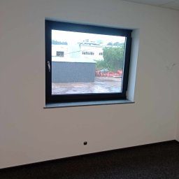 Andrzej Szelag - Wnętrze biura w trakcie wykańczania: świeżo pomalowane ściany na biało, czarna rama okienna z widokiem na zewnątrz w pochmurny dzień, ciemna wykładzina podłogowa.