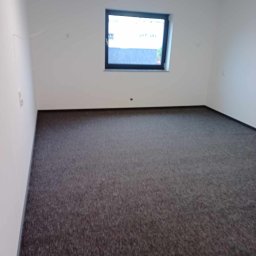 Andrzej Szelag - Pusta przestrzeń biurowa z szarą wykładziną dywanową, białymi ścianami i jednym oknem z czarną ramą. Widoczne ślady po demontażu na ścianie.