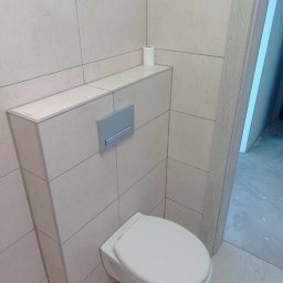 Andrzej Szelag - Nowoczesna łazienka z podwieszaną toaletą, jasnymi płytkami i szarym przyciskiem spłukującym; rolka papieru toaletowego na półce.