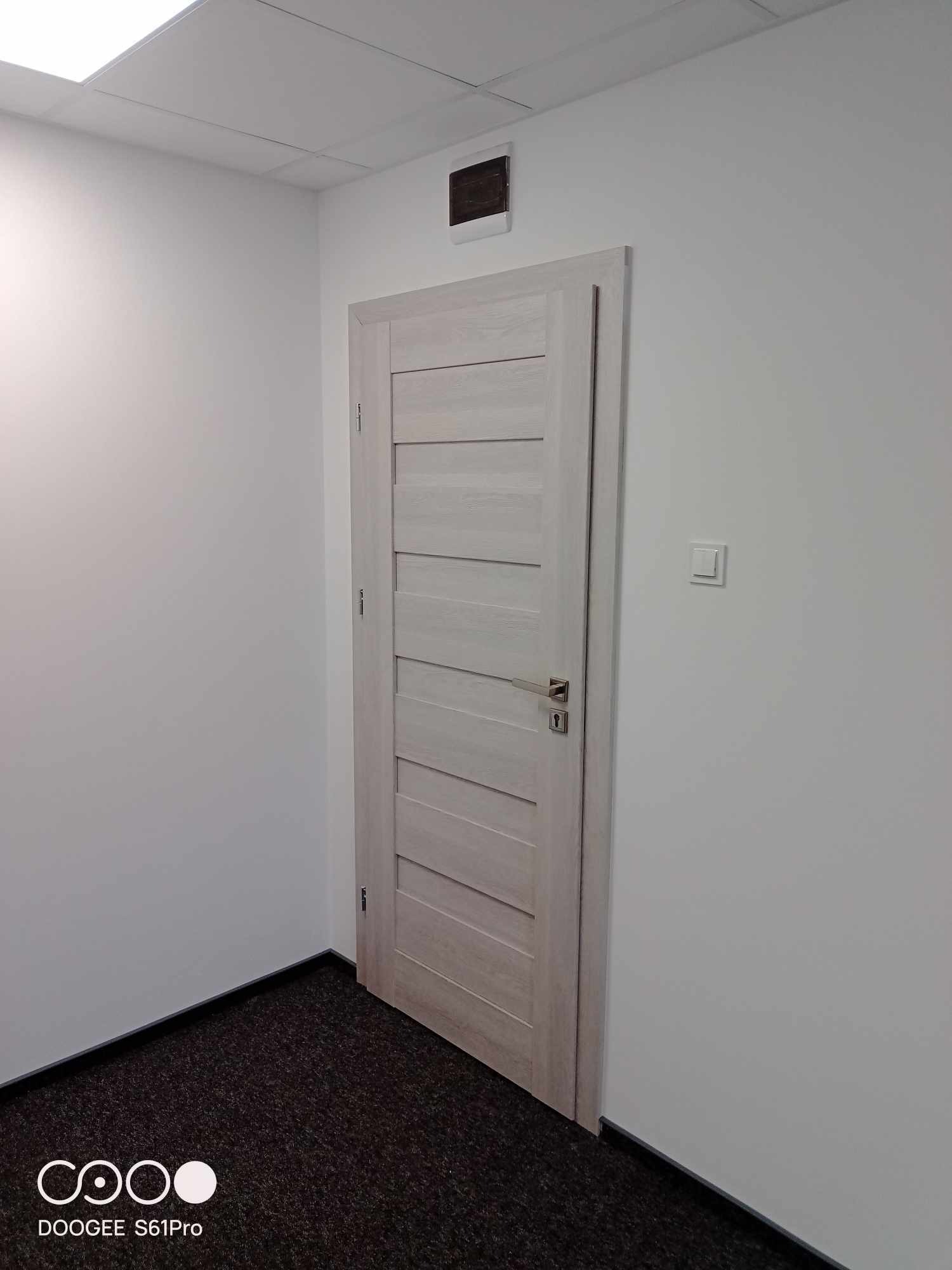 Wnętrze biura z jasnymi ścianami, ciemną wykładziną i drewnianymi drzwiami z poziomymi panelami, widoczny fragment sufitu podwieszanego i czujnik dymu.