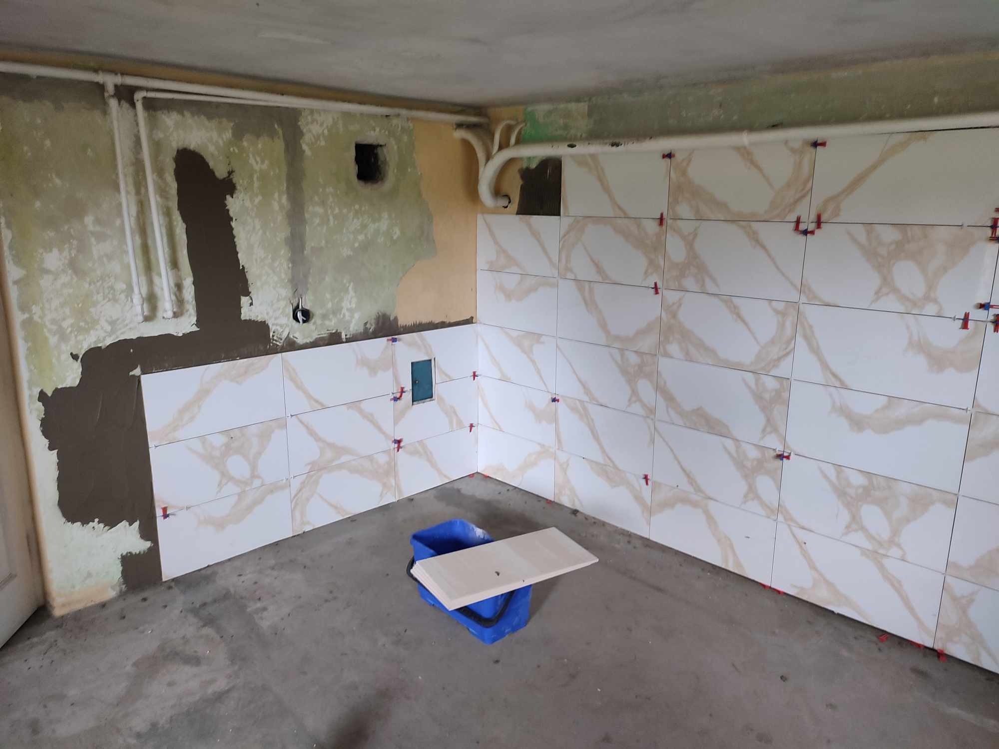 Ściana w trakcie układania płytek imitujących marmur, widoczne krzyżyki dystansowe, fragmenty nieotynkowanej ściany i wiadro z płytkami.