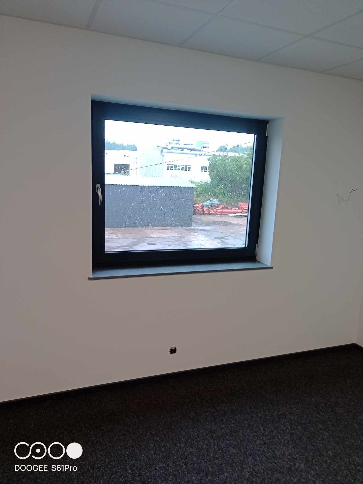 Wnętrze biura w trakcie wykańczania: świeżo pomalowane ściany na biało, czarna rama okienna z widokiem na zewnątrz w pochmurny dzień, ciemna wykładzina podłogowa.