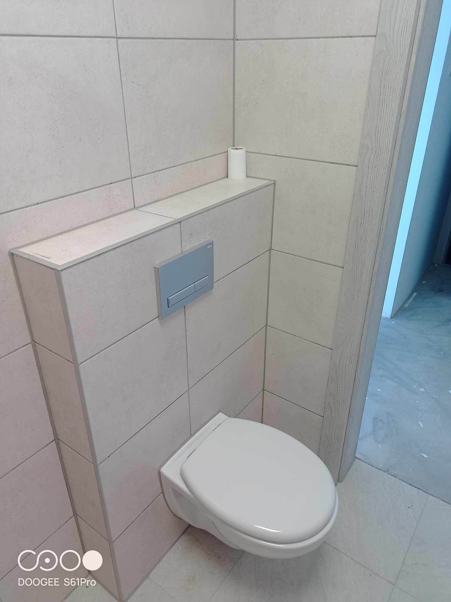 Nowoczesna łazienka z podwieszaną toaletą, jasnymi płytkami i szarym przyciskiem spłukującym; rolka papieru toaletowego na półce.